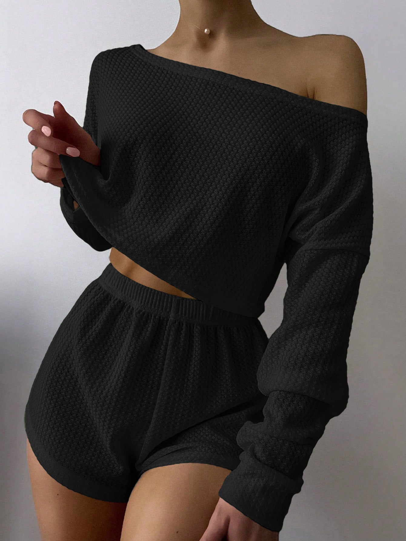 Woman wearing black off-shoulder knit two-piece loungewear set with crop top and shorts against white background