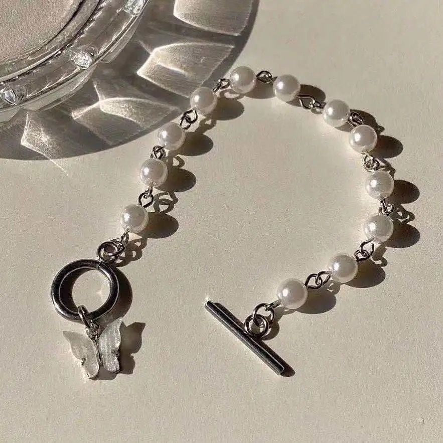 A pearl bracelet with silver links, toggle clasp and butterfly charm displayed on a cream surface next to glass decor