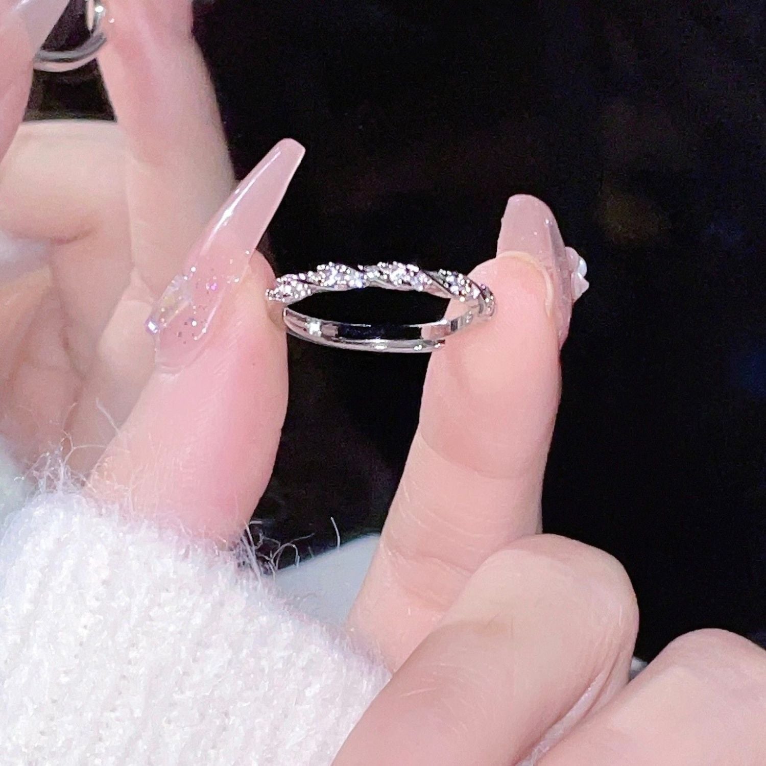 Hand with long pink nails holding delicate twisted silver ring with diamond accents against dark background