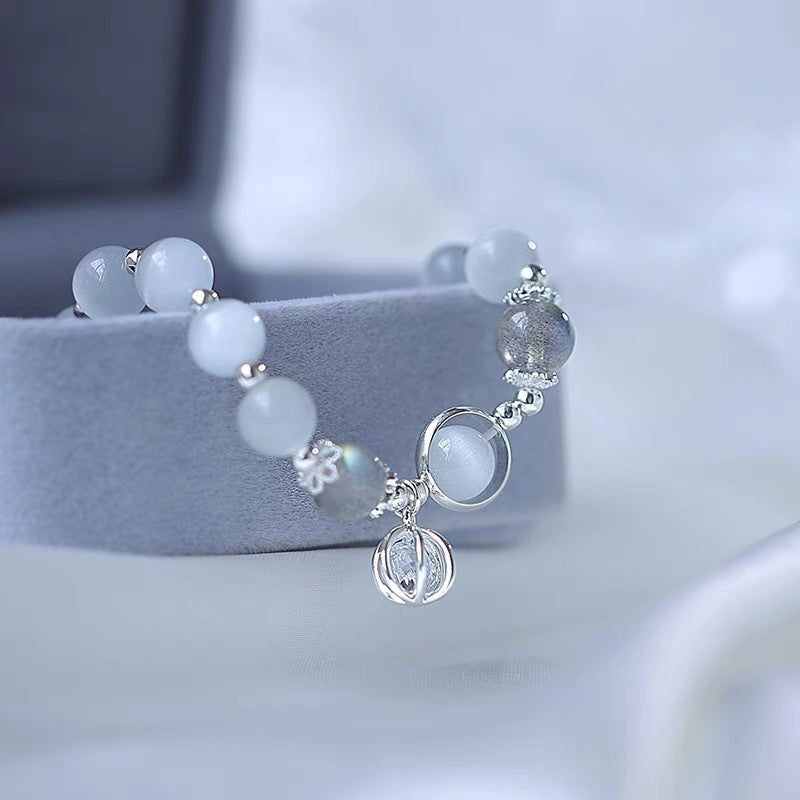 Delicate moonstone bracelet with silver accents and crystal charm pendant, displayed on grey cushion