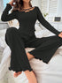 Woman wearing black ribbed loungewear set with criss-cross neckline and flared pants with ruffle hem, sitting casually on white bedding