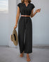 A woman wearing a black cap-sleeve jumpsuit with a tan belt, holding a wide-brimmed hat, standing against a neutral background.