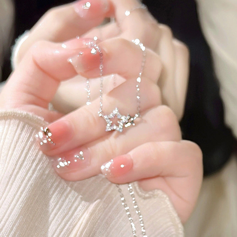 A woman is holding a delicate silver star pendant necklace with diamond accents in her palm, showing glitter nail art and cream sweater sleeve