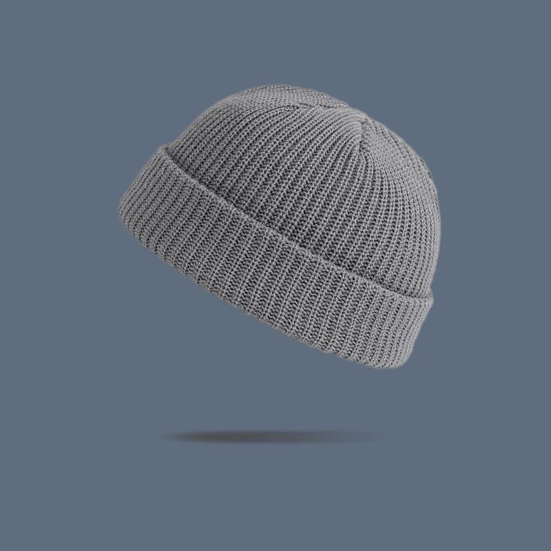 A light gray ribbed knit beanie with folded cuff floating against slate gray background, displaying classic fisherman cap design