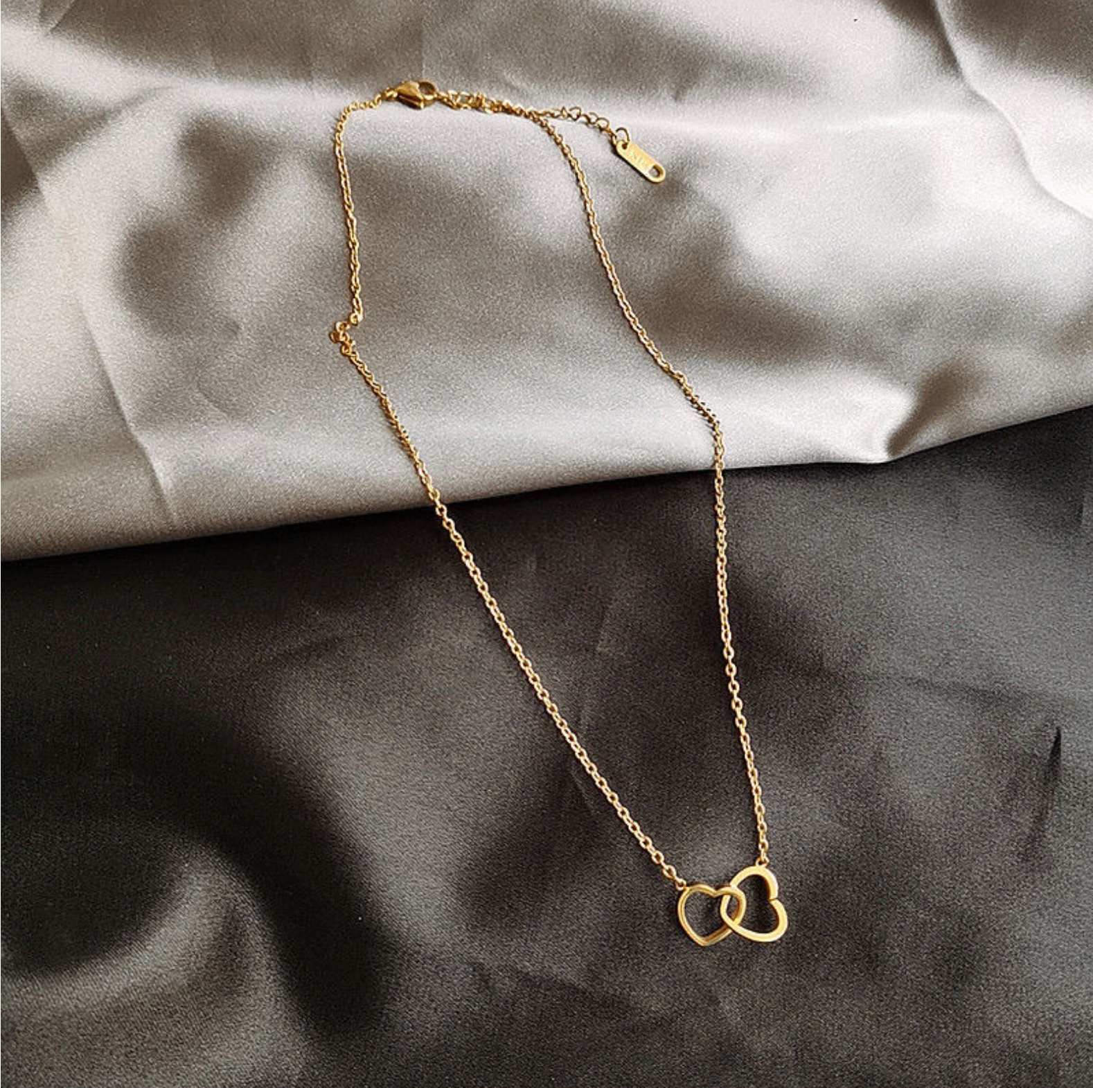 Gold chain necklace with interlocking heart pendant, laid flat on a gray fabric surface