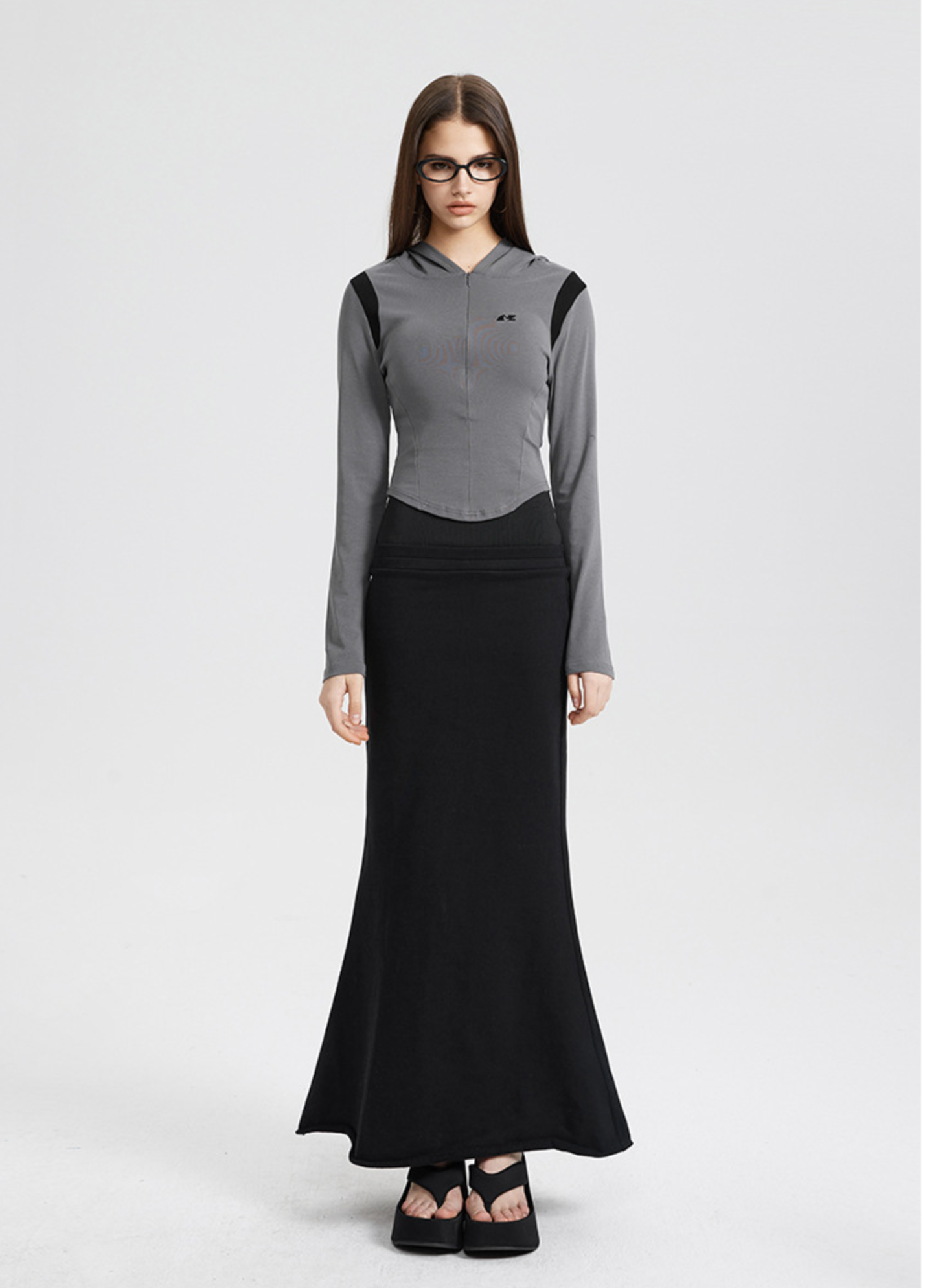 A woman is wearing a gray cropped hooded jacket with black accents and a black maxi skirt, standing straight against a white background