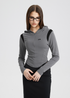 A woman is wearing black-framed glasses and a gray contoured cropped hoodie with black accents, standing against a white background