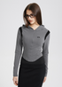 A woman is wearing glasses and a gray fitted hooded crop top with black shoulder panels, posing against a minimal white backdrop