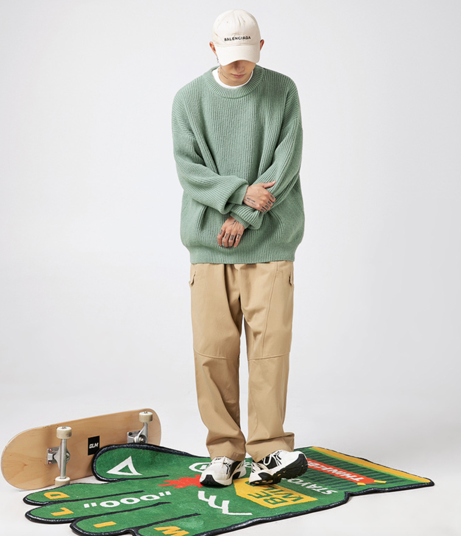 Full-length shot of sage green sweater with Balenciaga cap, khaki pants and white sneakers against white background