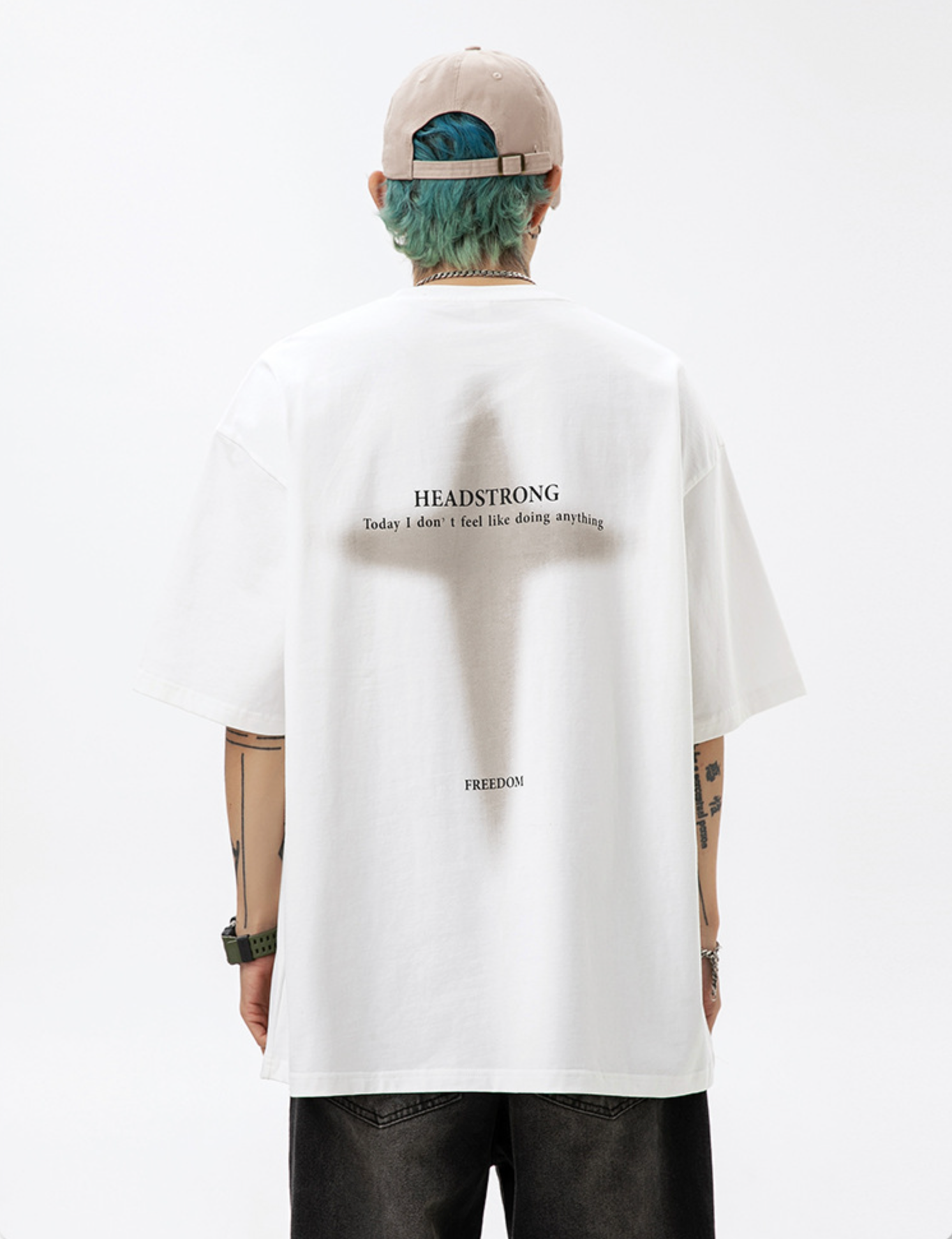 A man is standing with back to camera against white backdrop. Wearing white tee with cross graphic and "HEADSTRONG" text, beige cap and blue-green hair