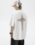 A man is standing in profile view against white background. Wearing white tee with cross graphic, white headphones, beige cap and showing arm tattoos