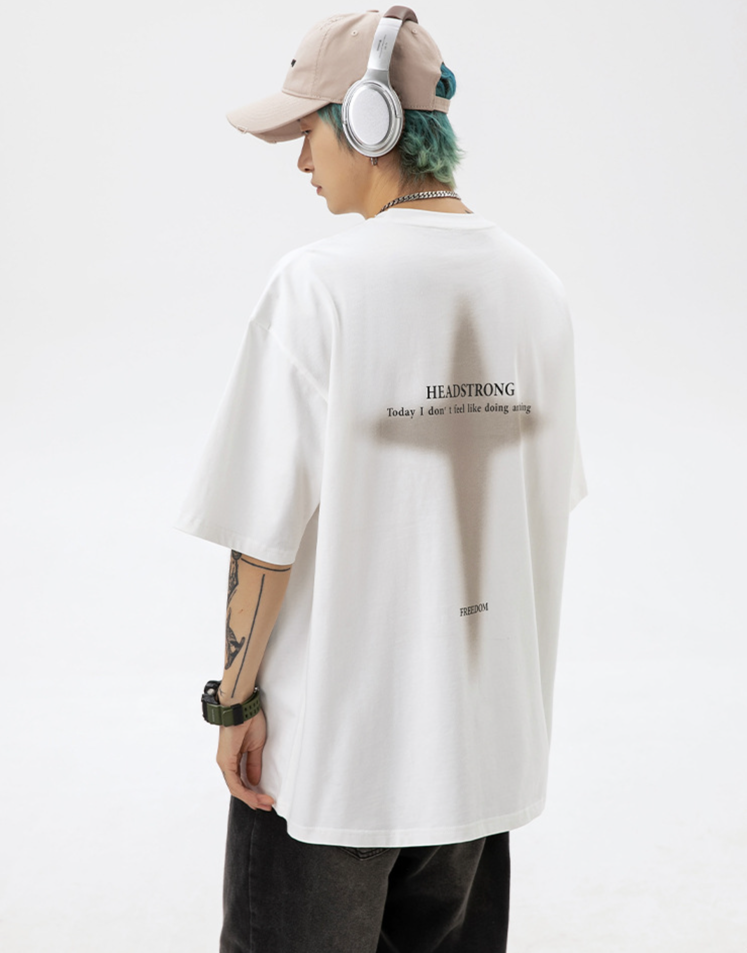 A man is standing in profile view against white background. Wearing white tee with cross graphic, white headphones, beige cap and showing arm tattoos