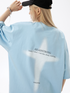 A woman is adjusting black cap with hand raised against white backdrop. Wearing baby blue oversized tee with white cross design and light blonde hair