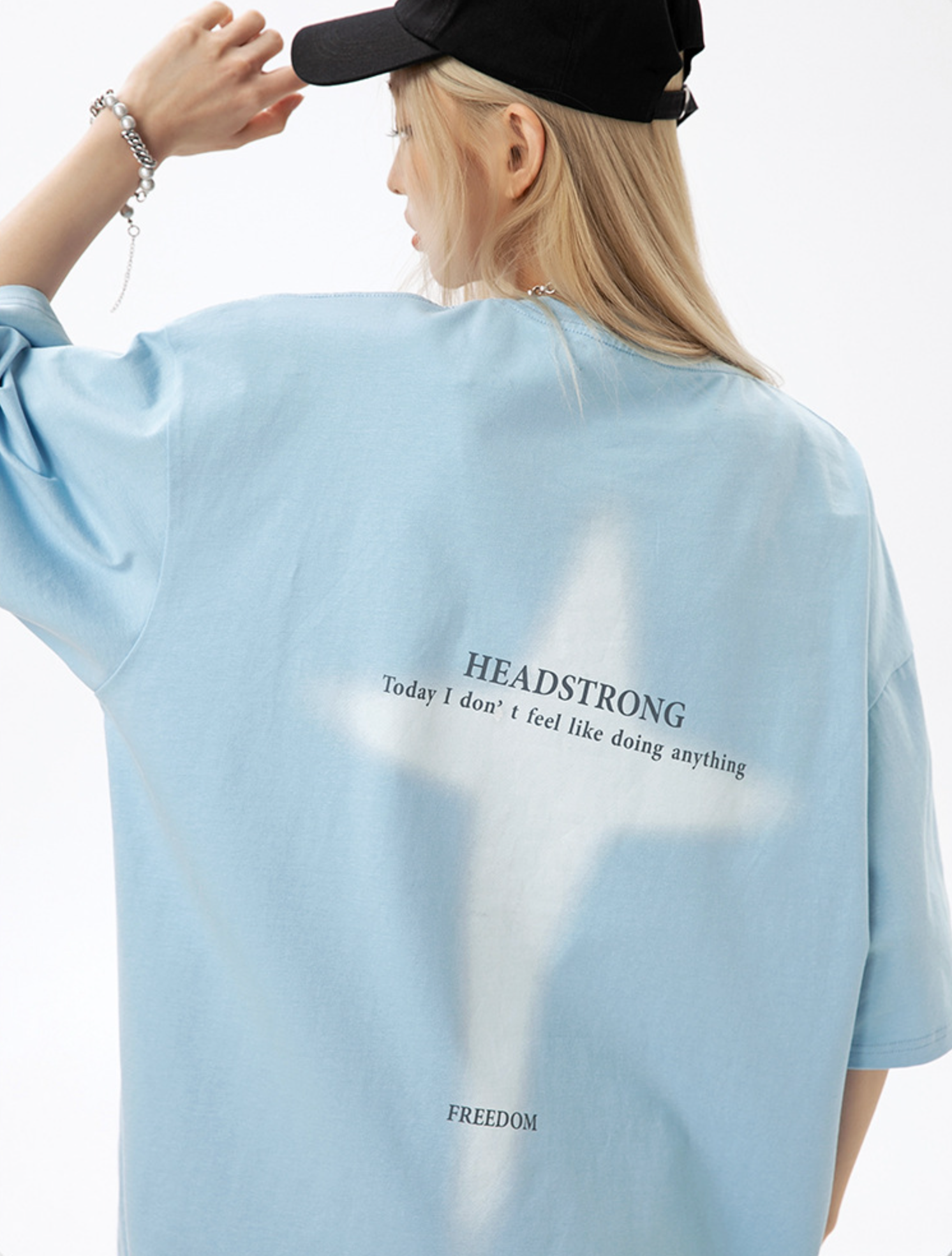 A woman is adjusting black cap with hand raised against white backdrop. Wearing baby blue oversized tee with white cross design and light blonde hair