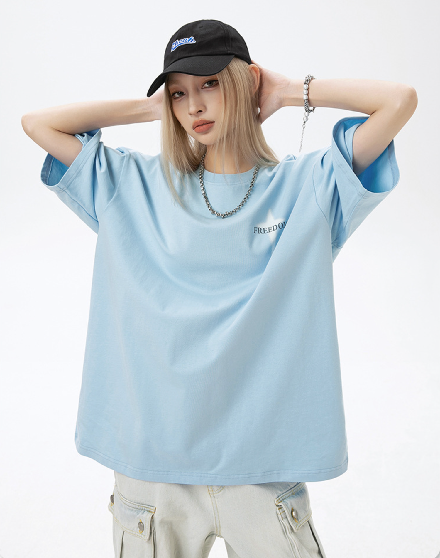 A woman is posing facing camera against white background. Wearing light blue tee with front FREEDOM text print, black cap and silver chain necklace