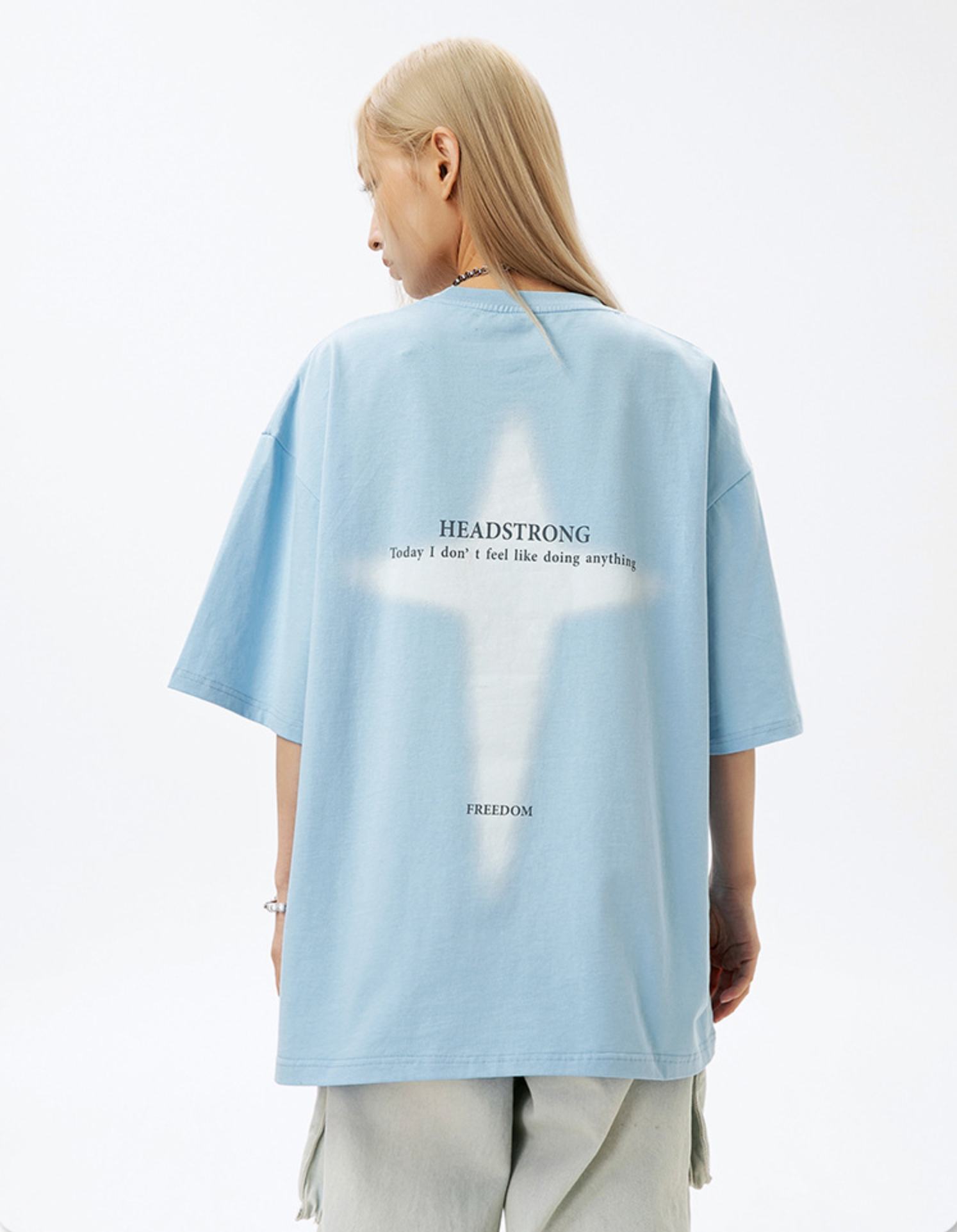 A woman is shown from behind against white backdrop. Wearing baby blue tee with white cross graphic print and "HEADSTRONG" text, blonde straight hair