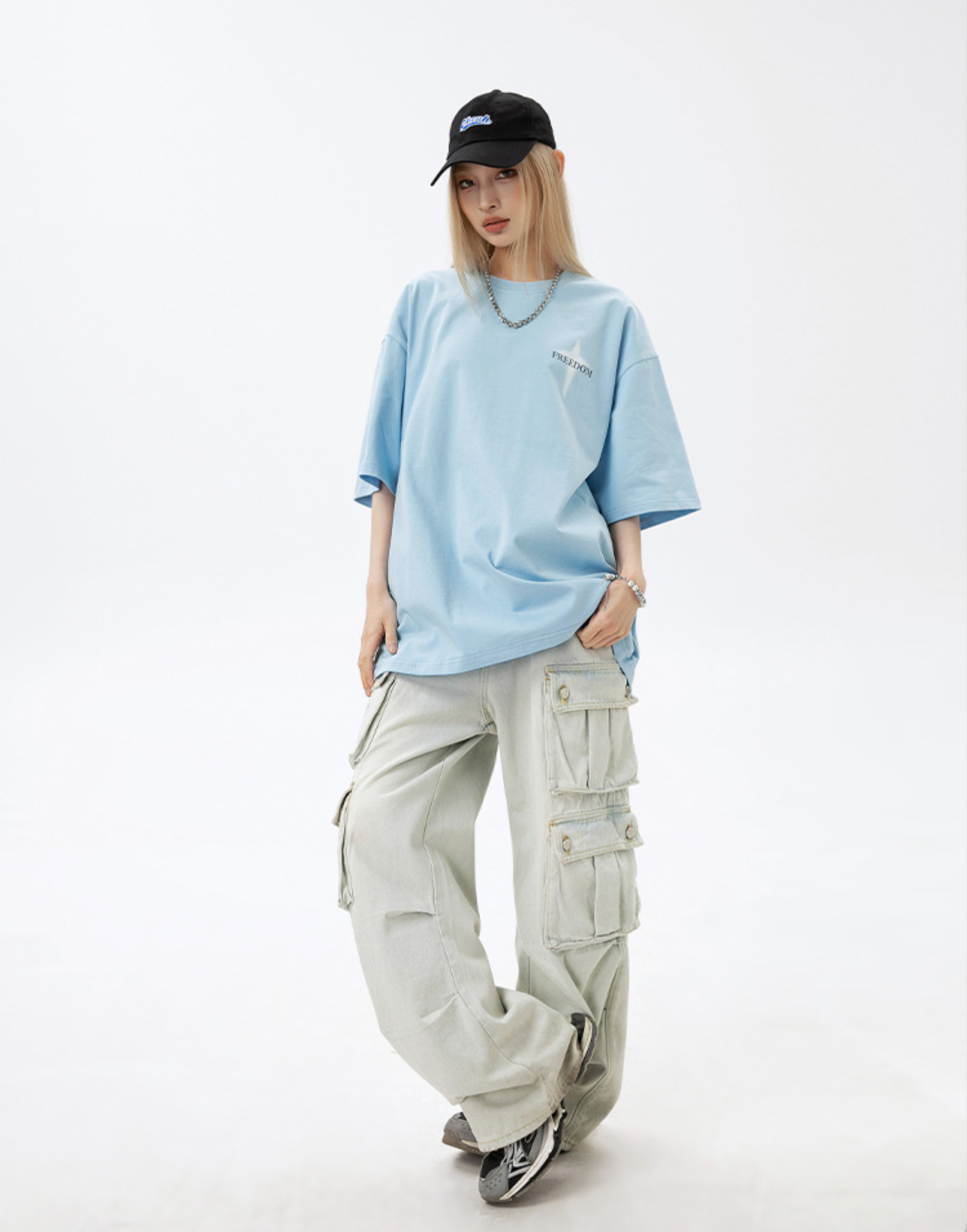 A woman is standing front-facing against white background. Wearing light blue oversized tee, black cap with blue logo and light beige cargo pants