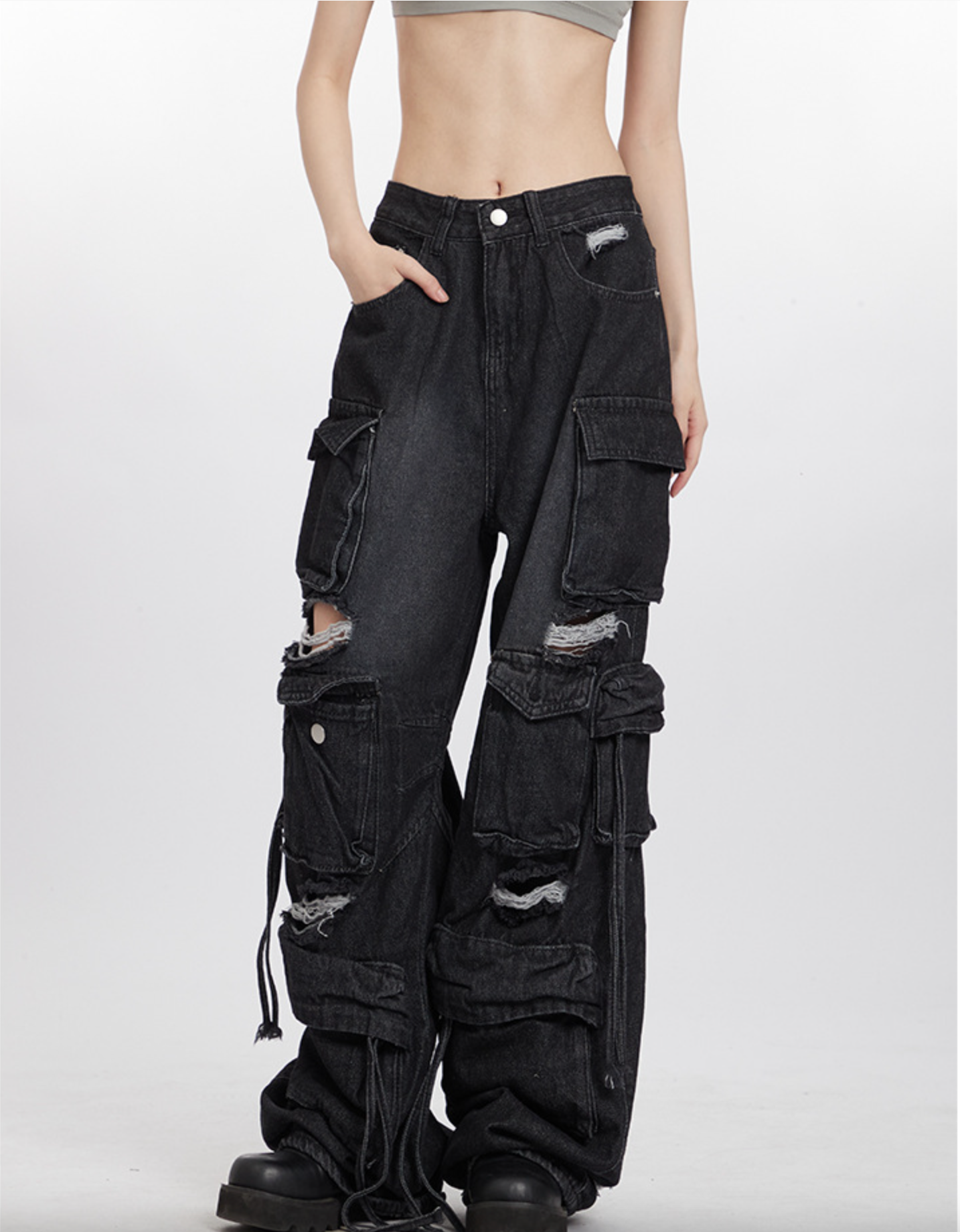 A model wearing grey crop top and black distressed cargo pants with multiple pockets, standing against white background