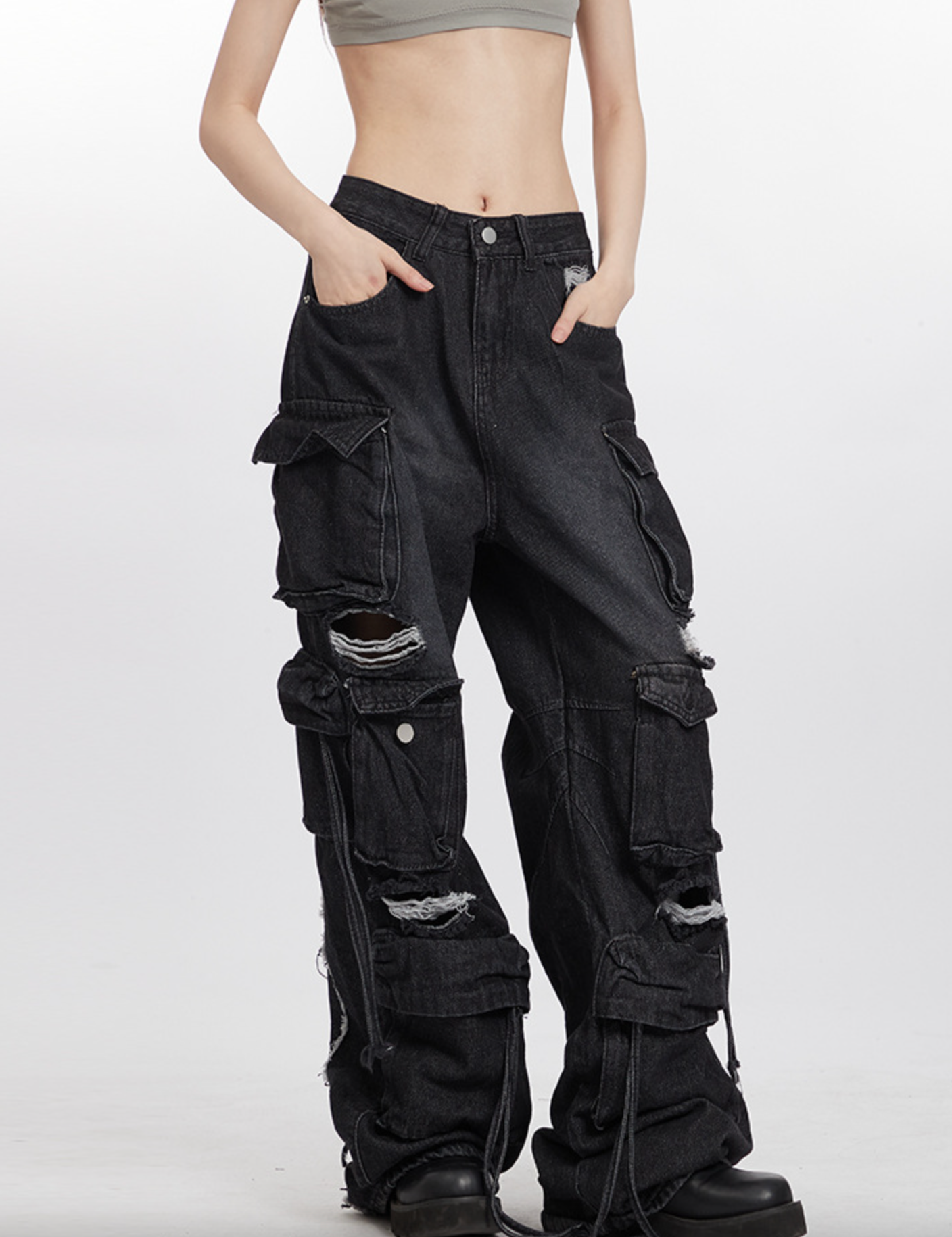 A model wearing grey crop top and black distressed cargo pants, showing front view with hands in pockets against white background
