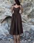 A model is posing in a chocolate brown linen sundress with fitted bodice against a rocky cliff background and pebble beach