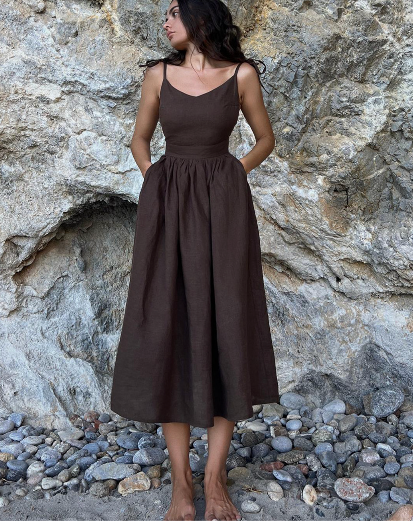 A model is posing in a chocolate brown linen sundress with fitted bodice against a rocky cliff background and pebble beach