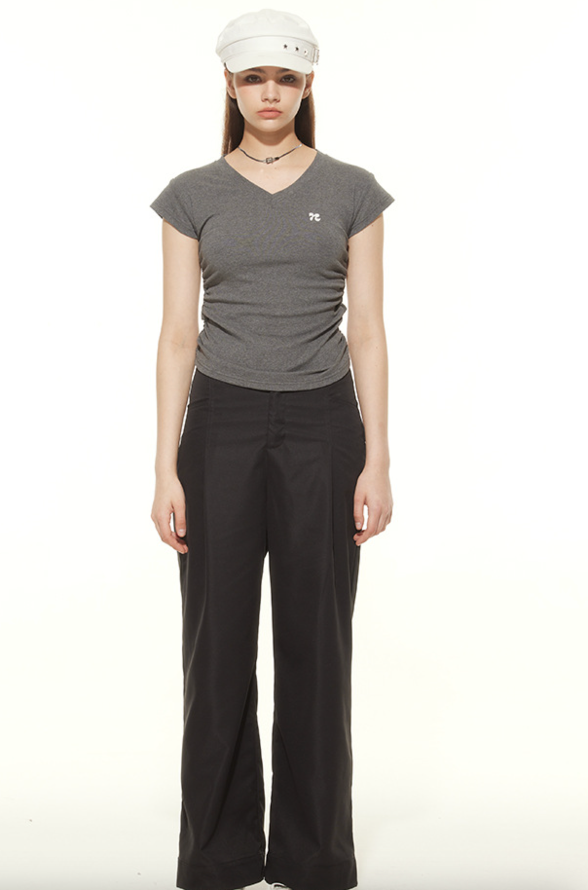 Woman wearing gray v-neck t-shirt with signature logo, black wide-leg pants and white military cap, full-length street style look