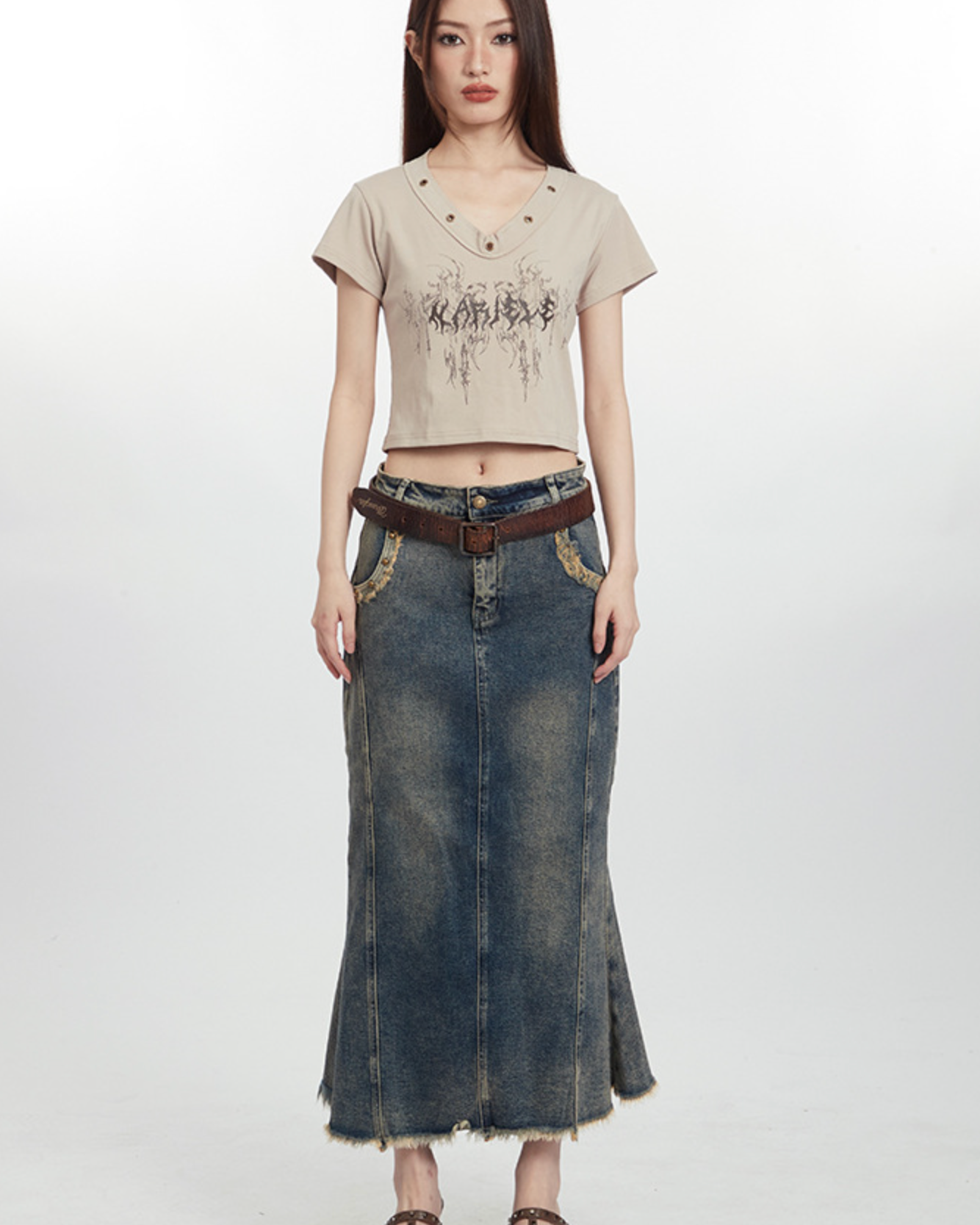Young woman wearing beige crop top with abstract print, long denim skirt, and brown leather belt against white background