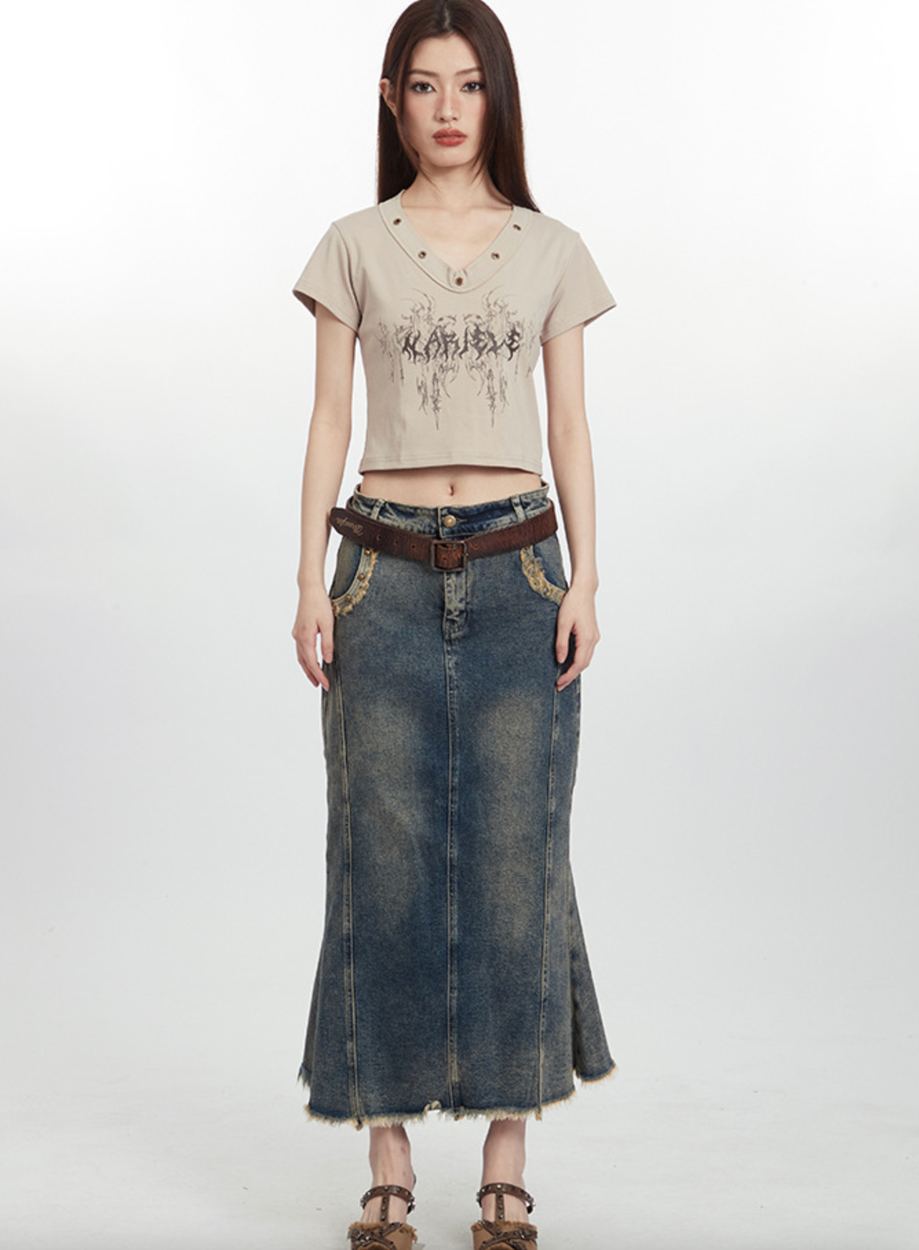 Young woman wearing beige crop top with abstract print, long denim skirt, and brown leather belt against white background