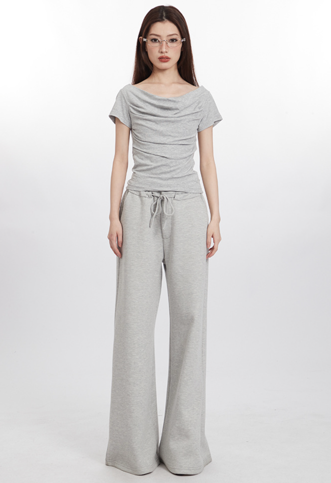 Full-body shot of model in light gray draped top and wide-leg pants, wearing glasses