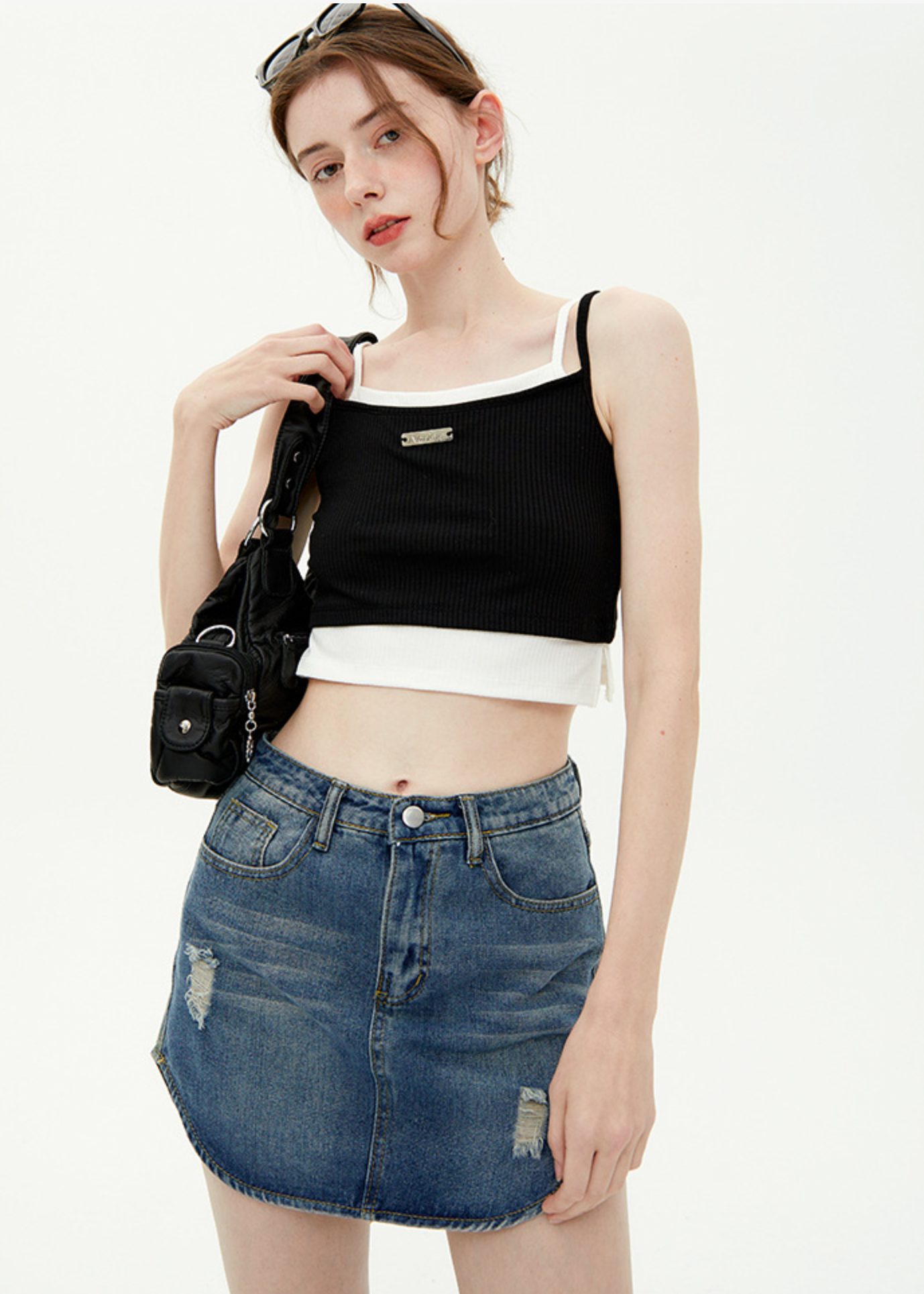 A woman is holding a small black bag while wearing a black ribbed crop top with white square neckline trim and distressed denim mini skirt