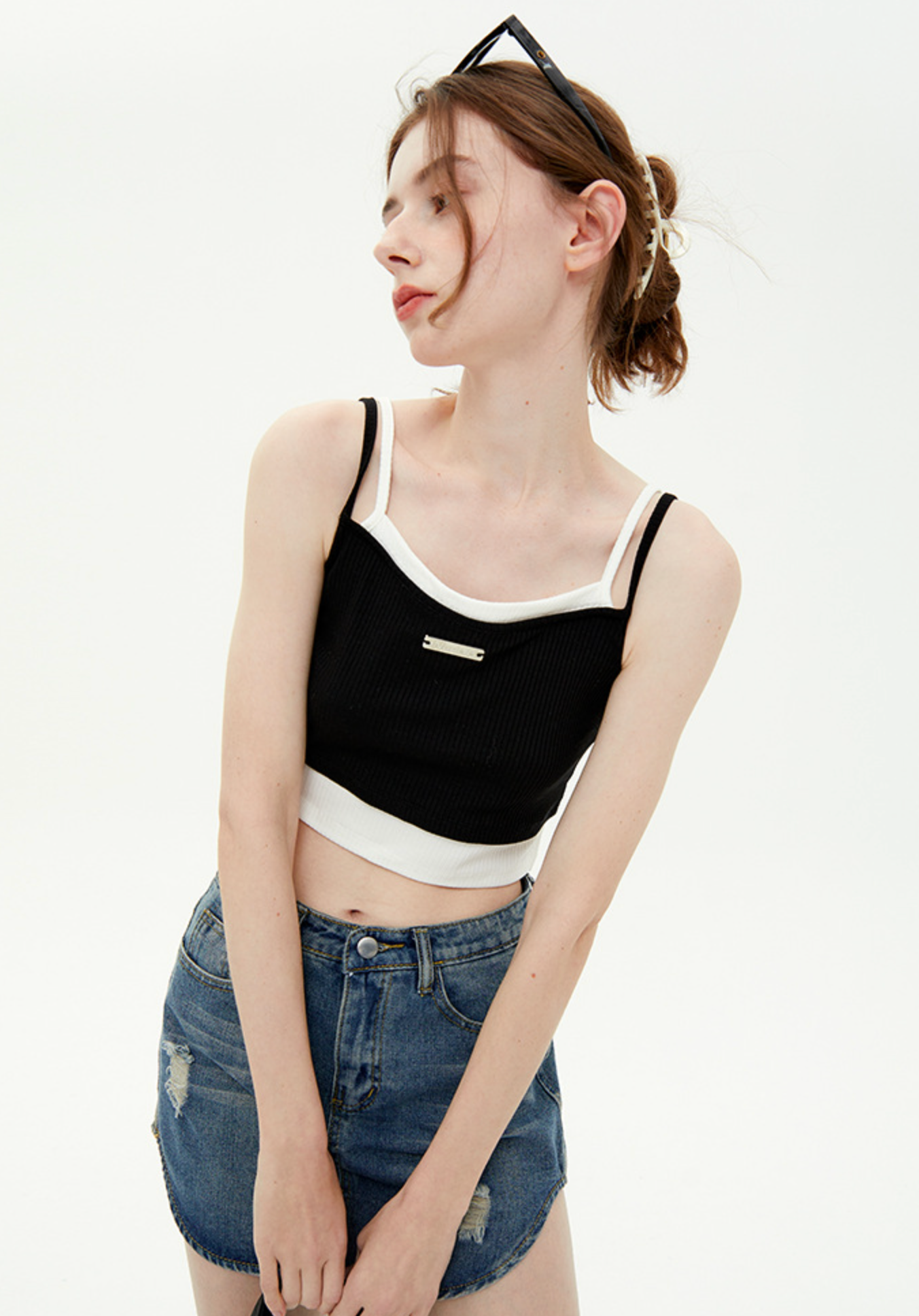 A woman is posing with head turned, wearing a black crop top with white trim detail, distressed denim mini skirt and sunglasses on her head