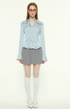 Woman in light blue blouse and pleated skirt, wearing glasses, white tights, white shoes