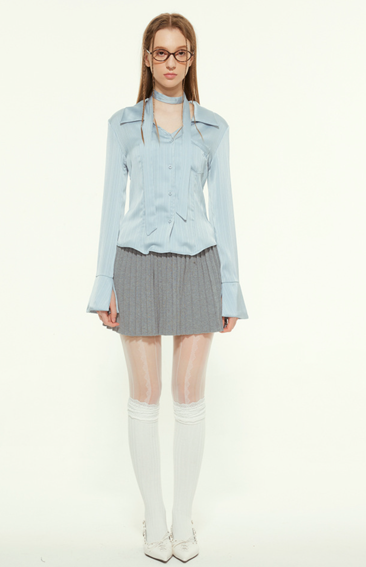 Woman in light blue blouse and pleated skirt, wearing glasses, white tights, white shoes