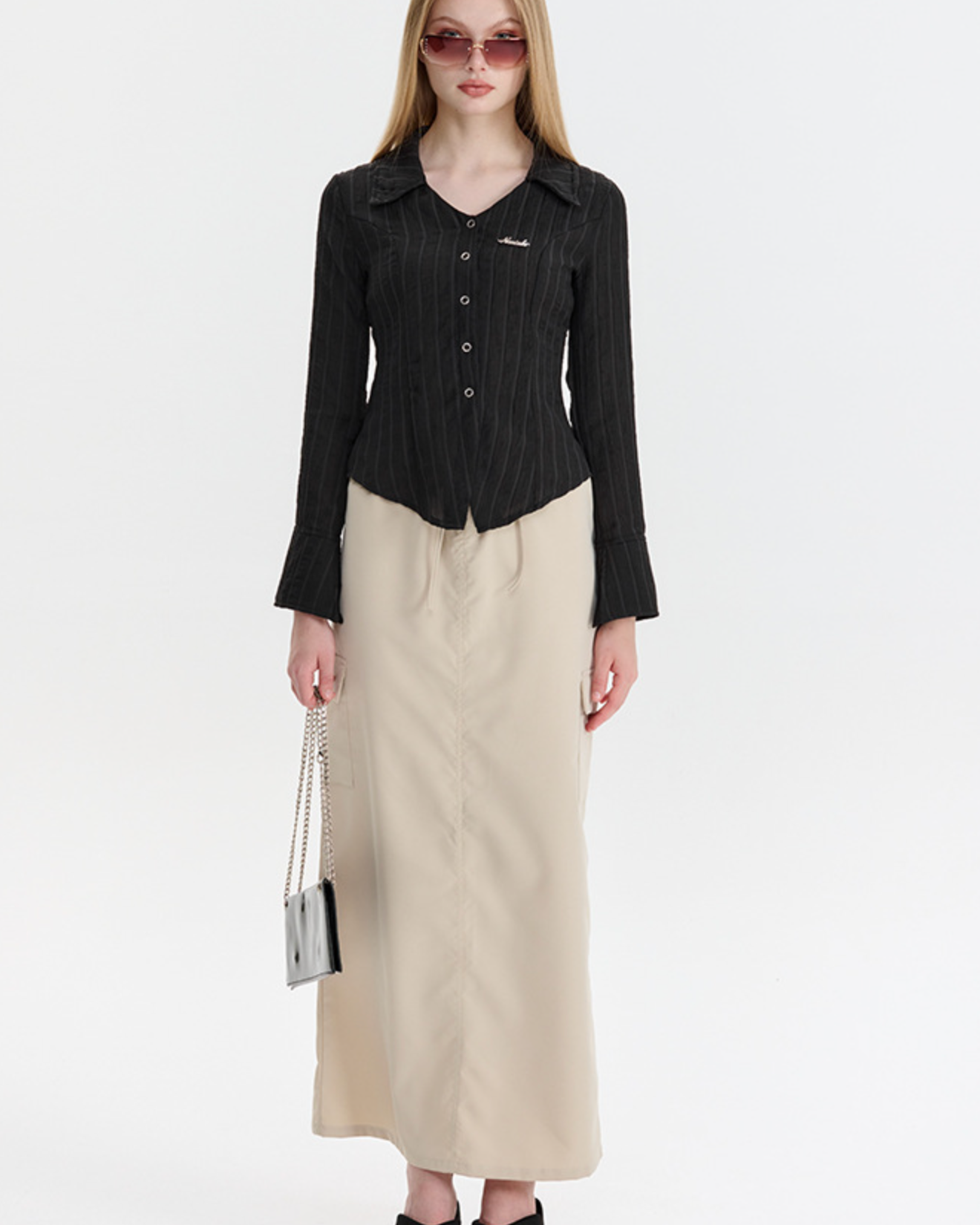 A woman is posing in a black pinstriped button-up shirt with bell sleeves and beige maxi skirt, accessorized with sunglasses and small handbag