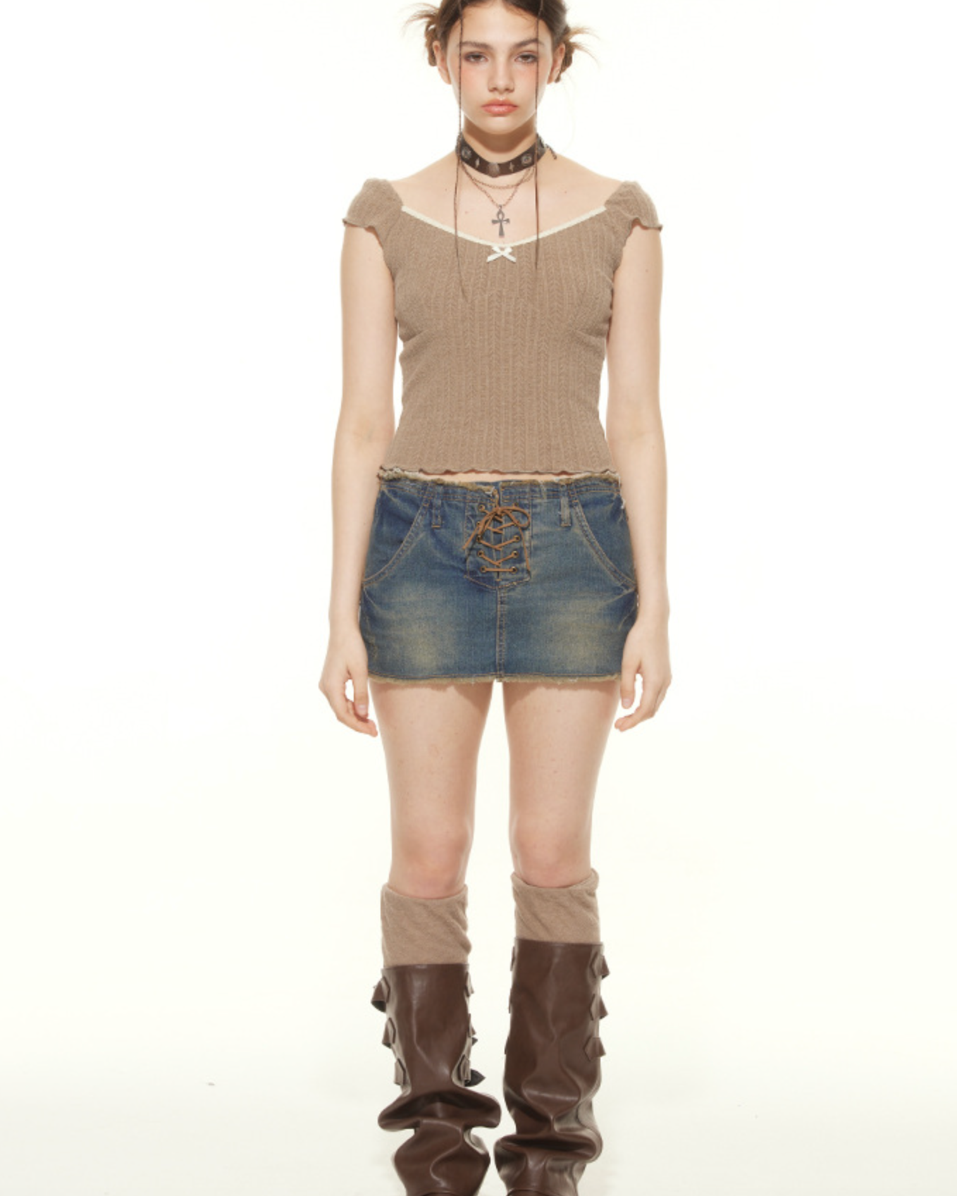 A woman wearing a tan ribbed crop top with white trim and lace-up denim mini skirt stands with brown knee-high boots against white background
