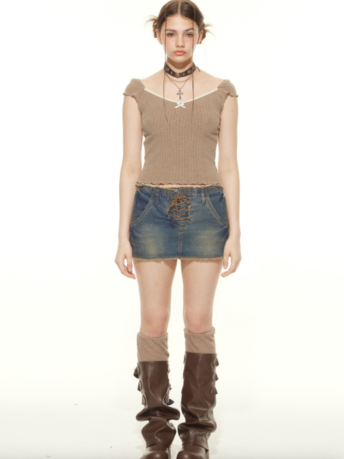 A woman wearing a tan ribbed crop top with white trim and lace-up denim mini skirt stands with brown knee-high boots against white background