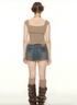 A woman wearing a tan ribbed crop top with white trim poses from behind, showing raw-edge denim mini skirt and brown buckled boots