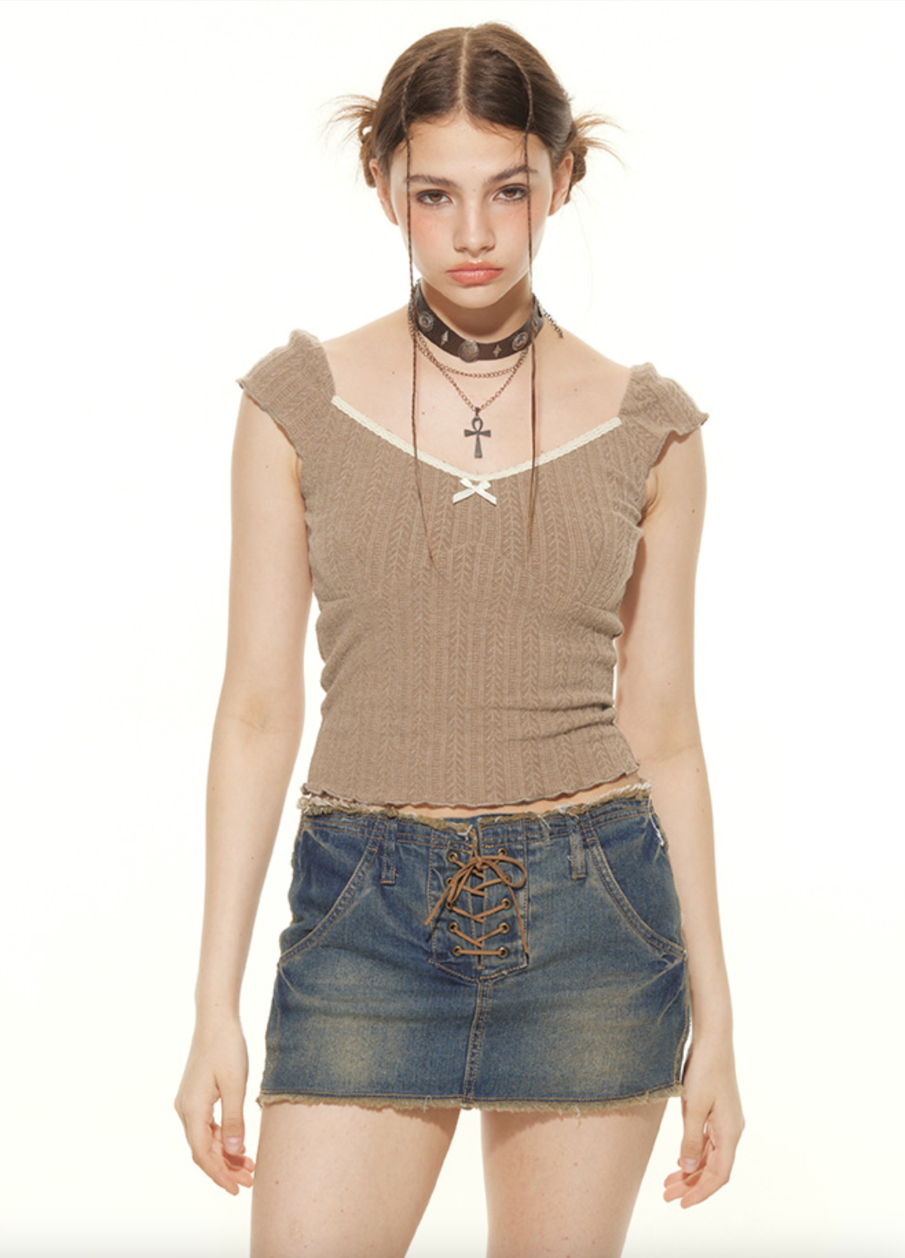 A woman wearing a tan cable-knit crop top with studded choker stands facing camera in lace-up denim mini skirt against white background