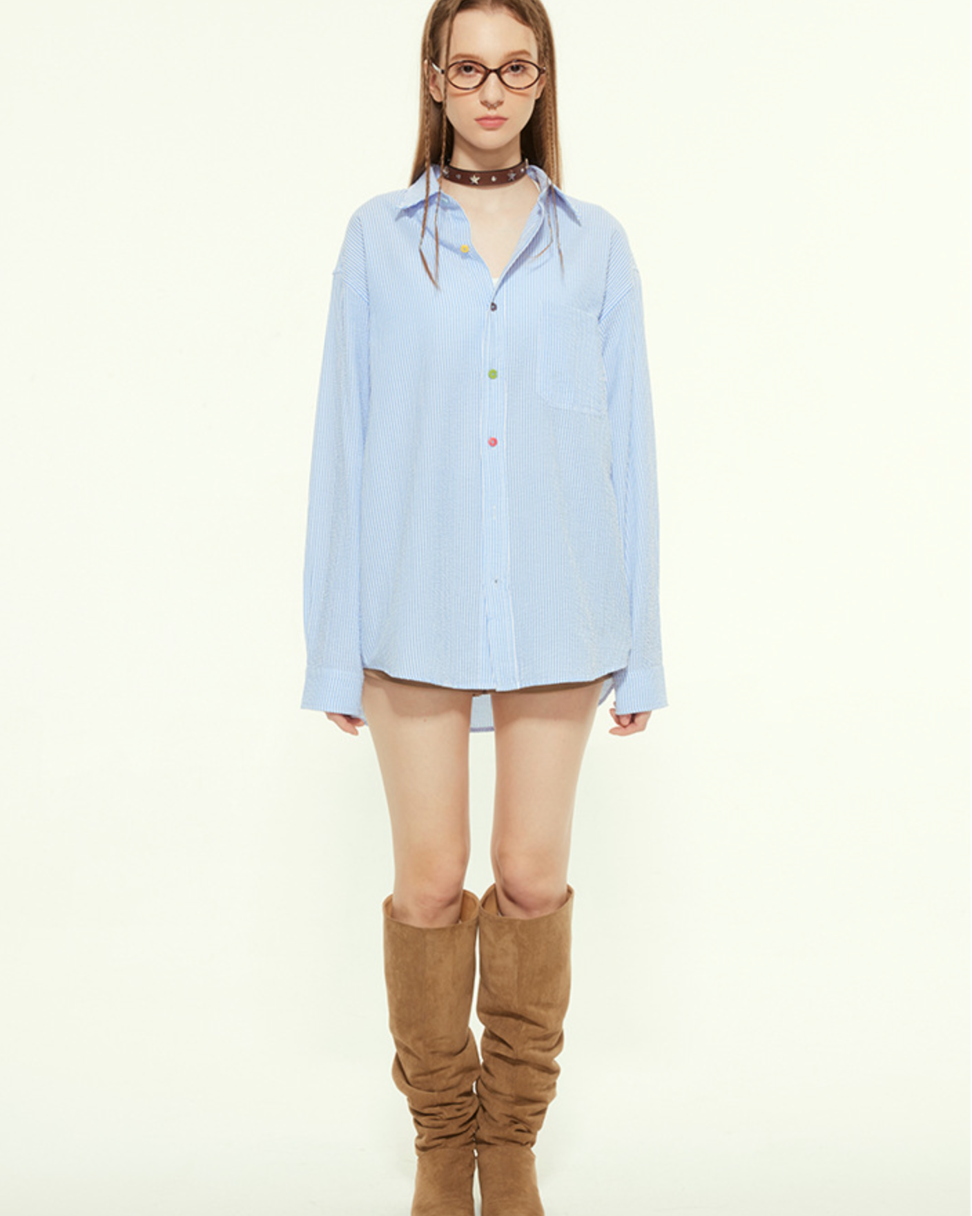 A female model is wearing an oversized blue striped button-up shirt with multicolored buttons and knee-high brown boots, standing against a plain background