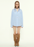A female model is wearing an oversized blue striped button-up shirt with multicolored buttons and knee-high brown boots, standing against a plain background