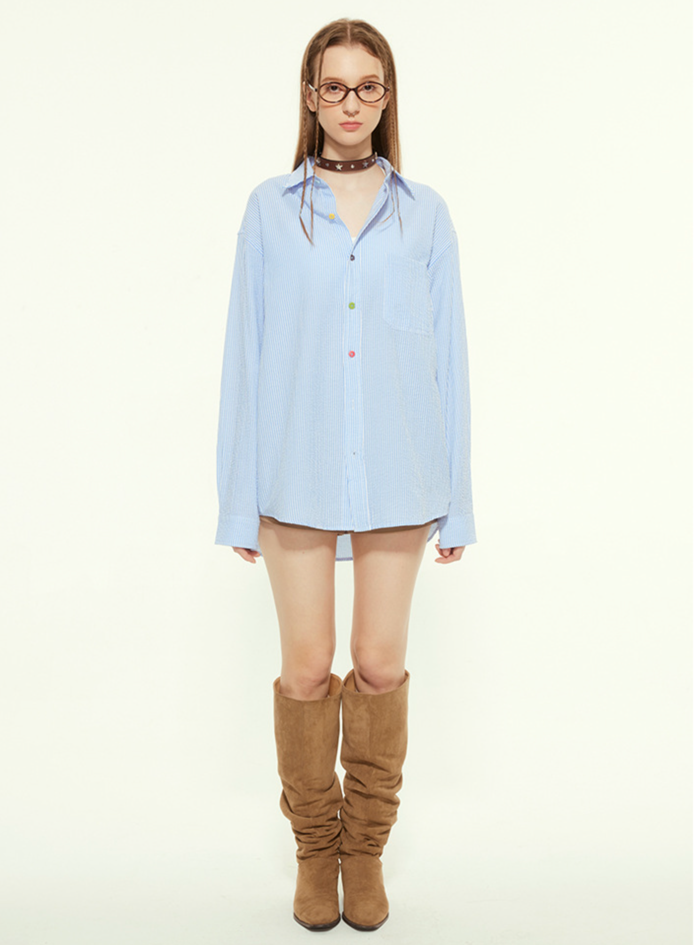 A female model is wearing an oversized blue striped button-up shirt with multicolored buttons and knee-high brown boots, standing against a plain background