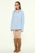 A female model is wearing a blue striped button-up shirt with multicolored buttons, standing against a neutral background