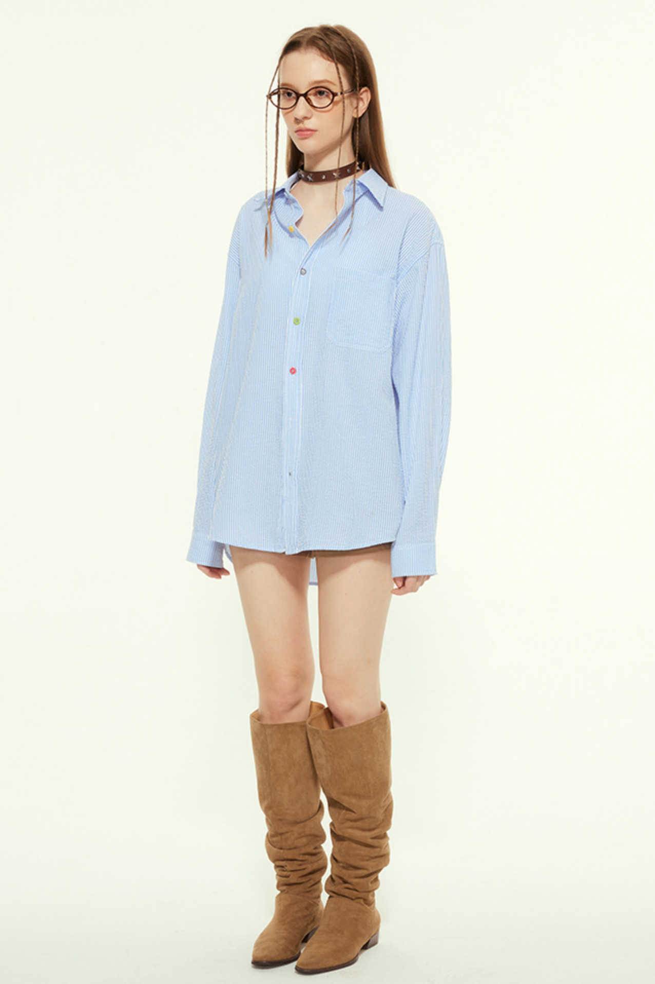 A female model is wearing a blue striped button-up shirt with multicolored buttons, standing against a neutral background