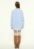 A female model is shown from the back wearing a loose blue striped button-up shirt with a white label on the upper back, paired with slouchy brown boots