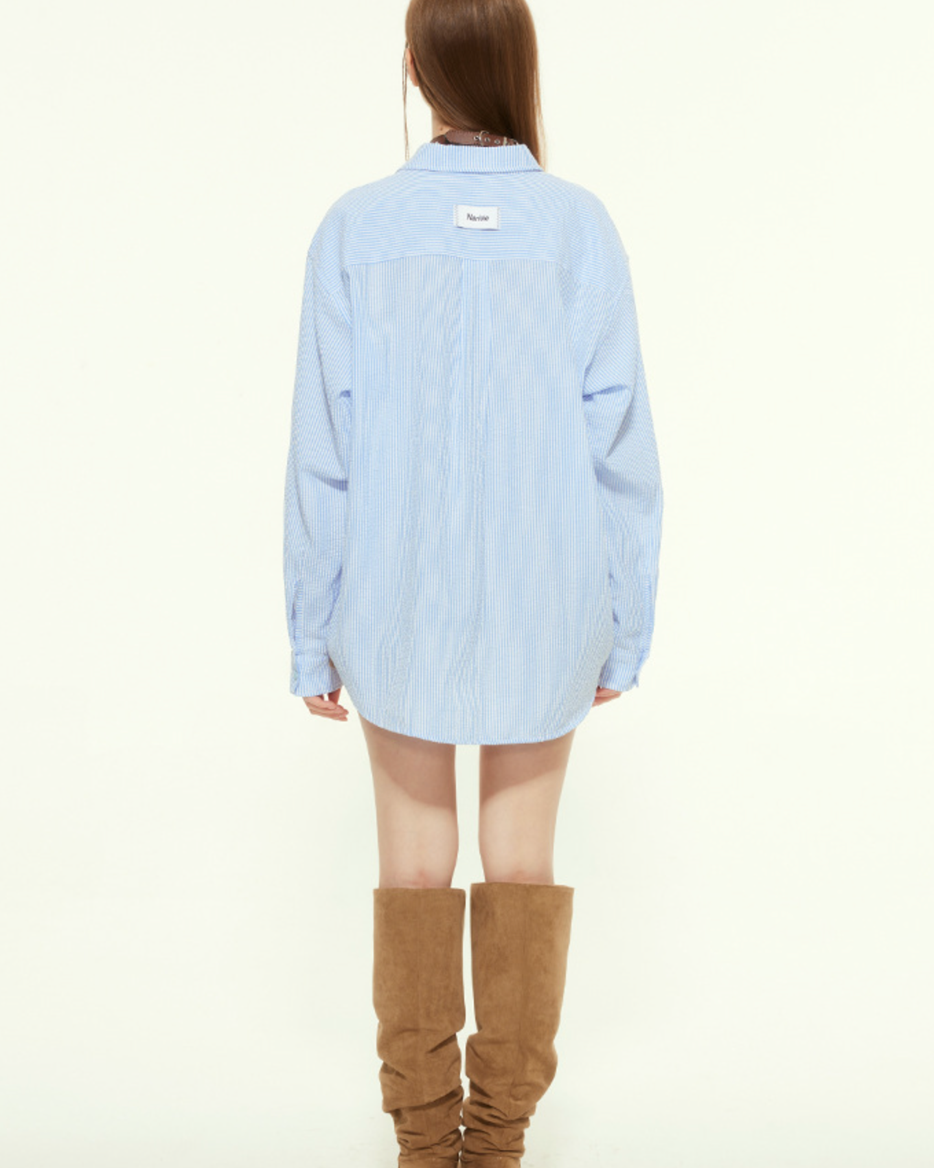 A female model is shown from the back wearing a loose blue striped button-up shirt with a white label on the upper back, paired with slouchy brown boots