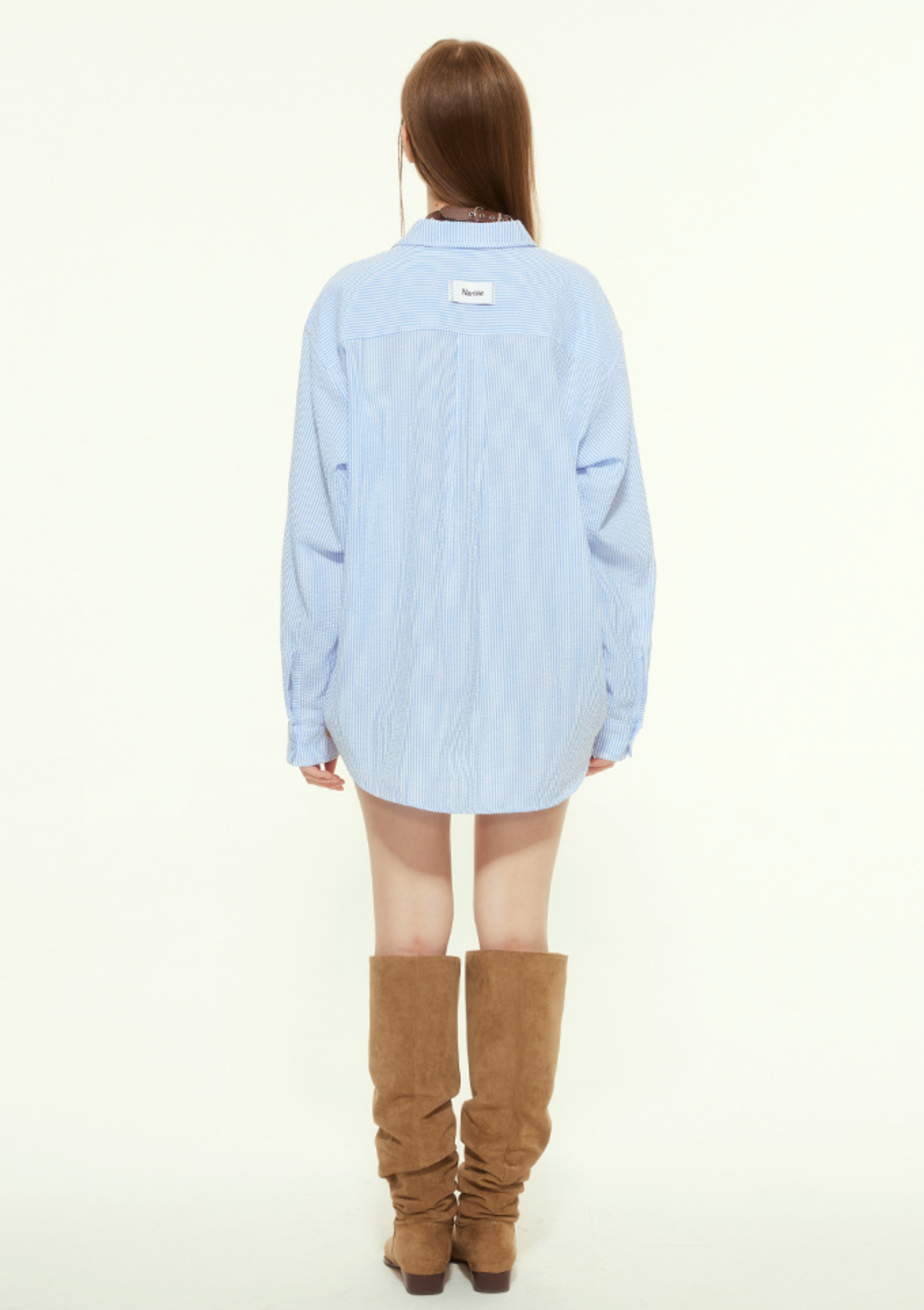 A female model is shown from the back wearing a loose blue striped button-up shirt with a white label on the upper back, paired with slouchy brown boots