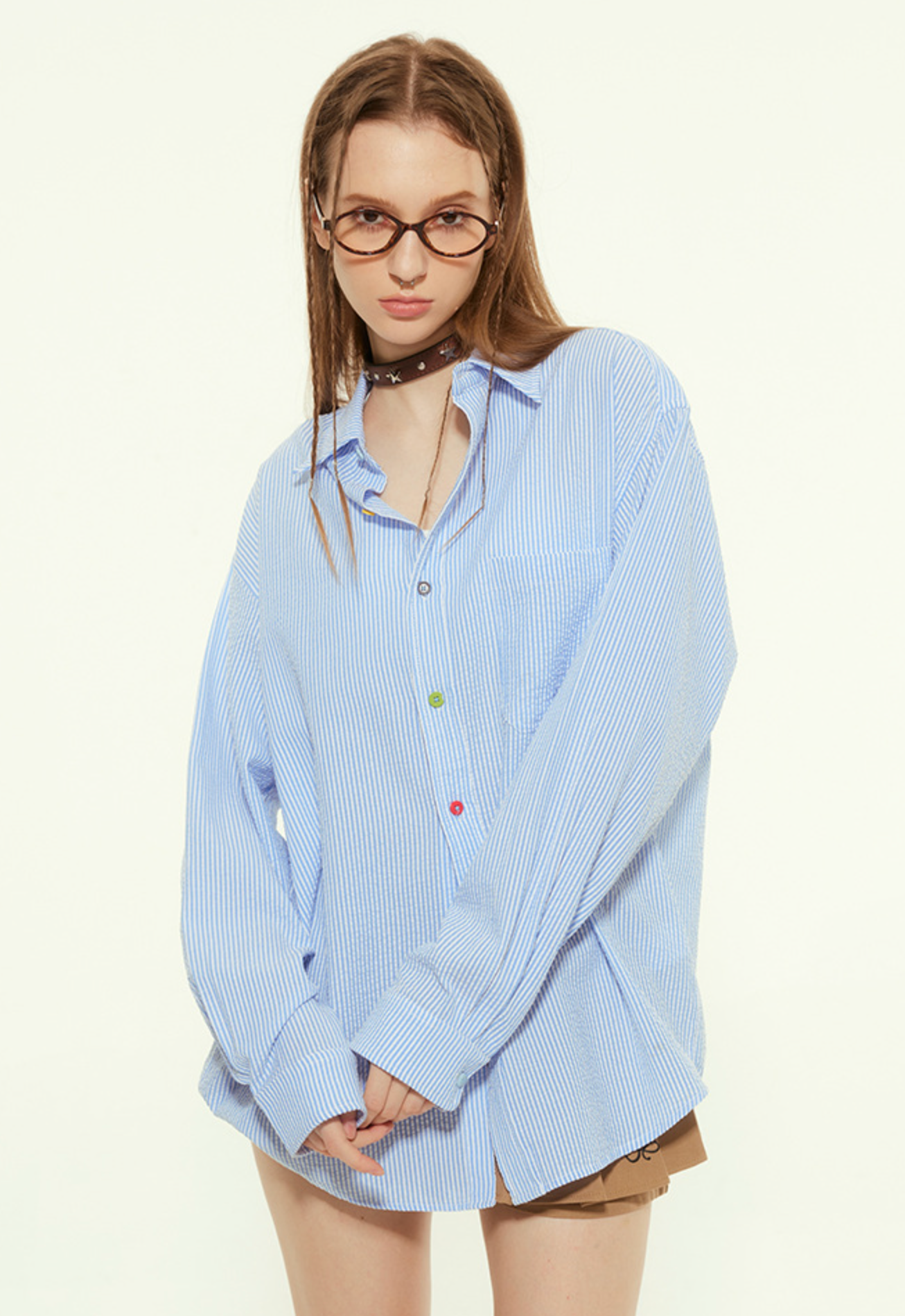 A female model is wearing a loose blue striped button-up shirt with a relaxed fit, styled with a choker and glasses, posing with slightly tucked hands