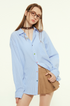 A female model is wearing a blue striped button-up shirt and a brown pleated mini skirt, posing with one hand on her hip and looking directly at the camera