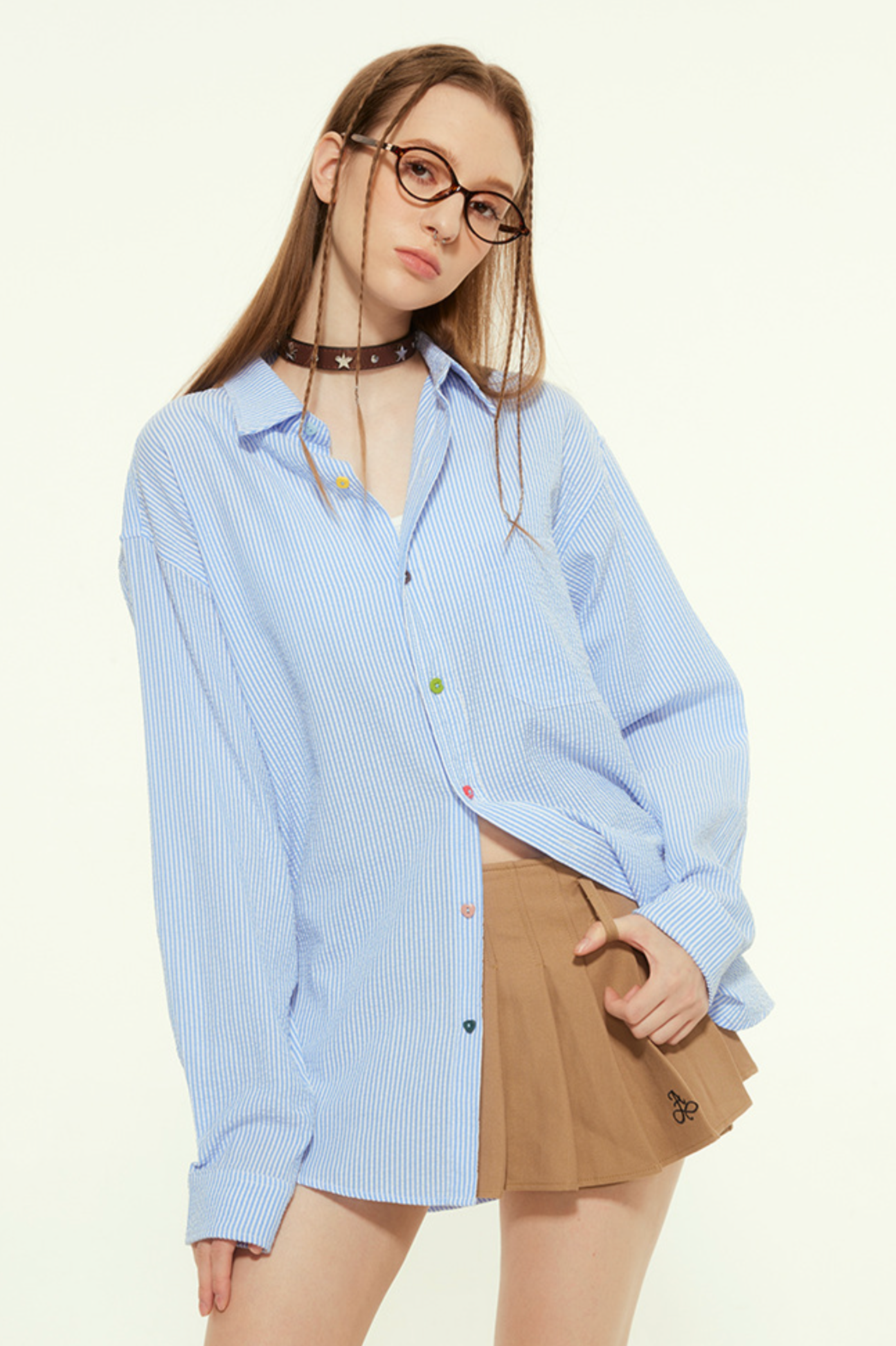 A female model is wearing a blue striped button-up shirt and a brown pleated mini skirt, posing with one hand on her hip and looking directly at the camera