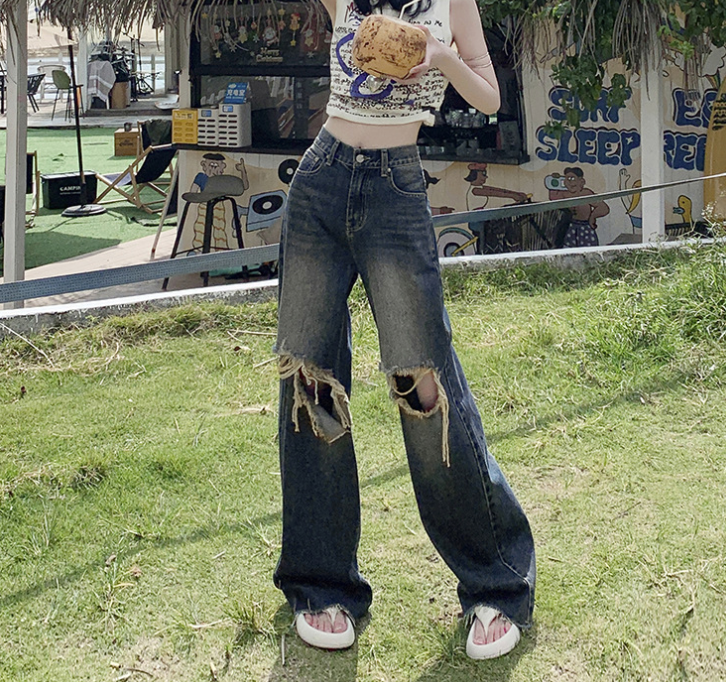 Woman wearing distressed wide-leg jeans with knee rips and white sandals, standing on grass near outdoor cafe
